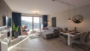 Ein modernes Wohnzimmer mit einer Couch, einem Esstisch, Stühlen, einem Fernseher und einem großen Fenster mit Vorhängen.