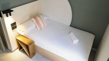 A neatly made bed with white bedding and a single pillow, a wooden bedside table, and a wall-mounted light fixture.