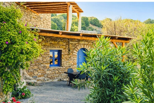 Un edificio de piedra con una pérgola de madera, una puerta azul y una mesa con sillas en el patio.