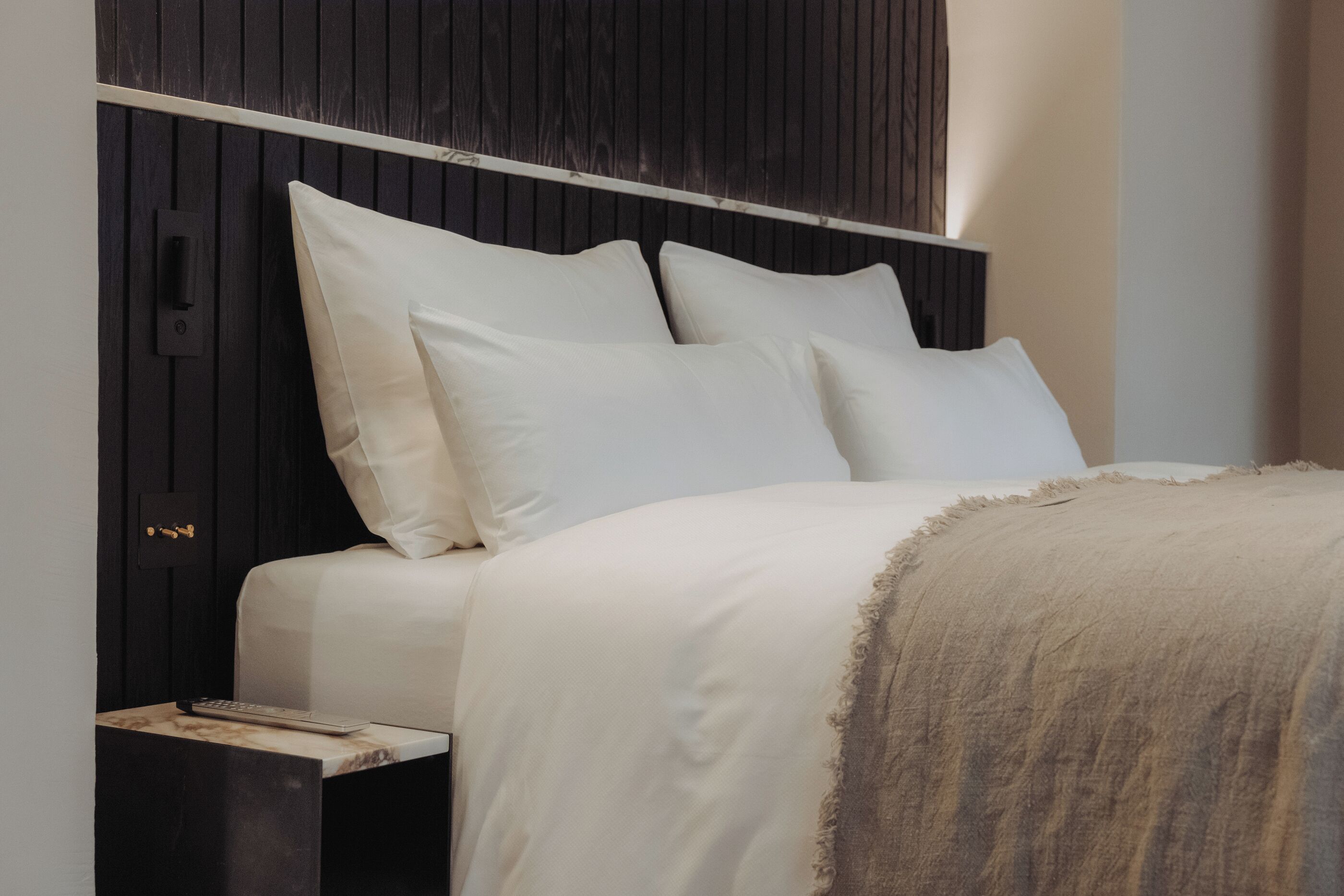 Una cama bien tendida con ropa blanca y una manta color beige, frente a un cabecero de madera oscura.