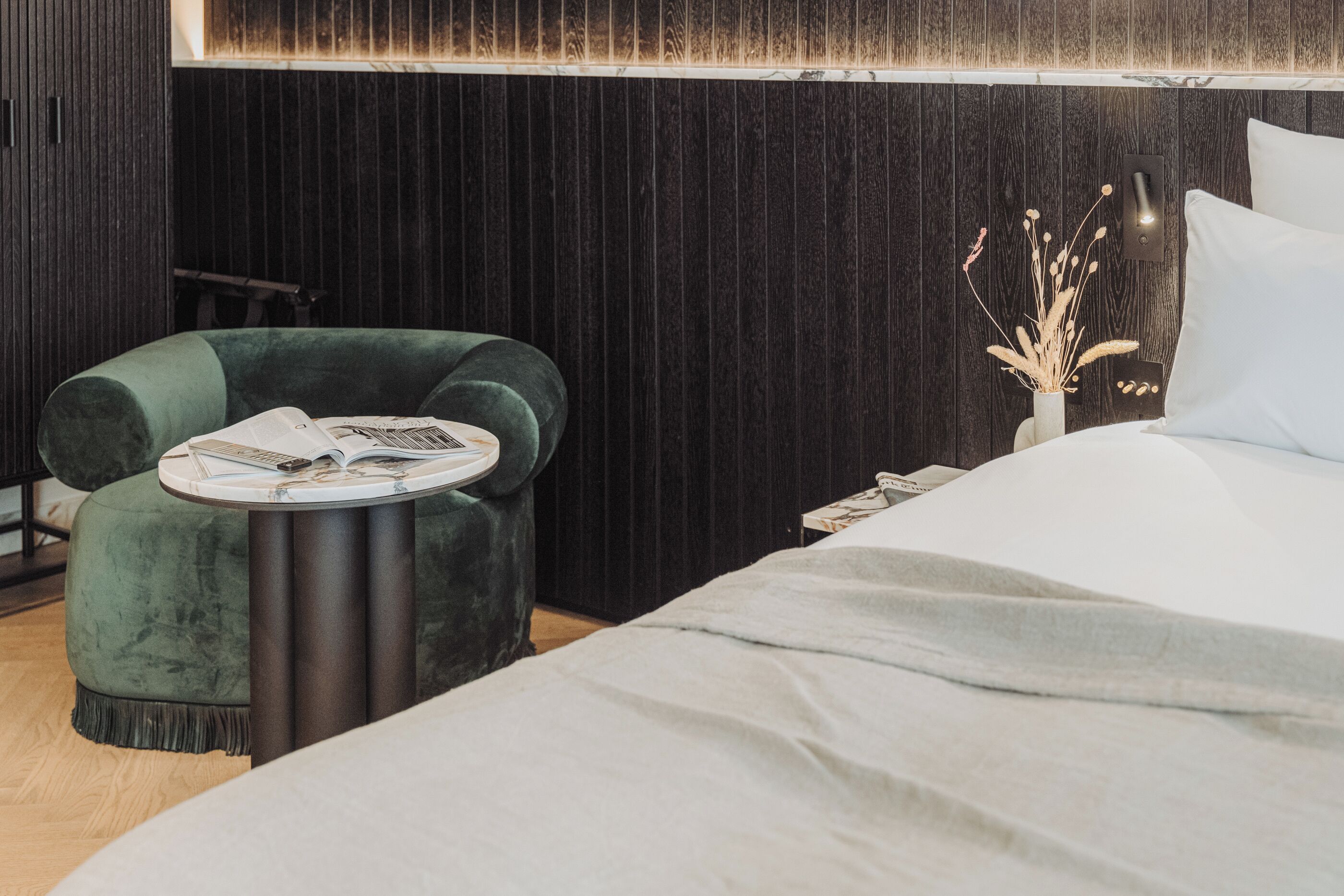 Une chambre moderne avec un fauteuil en velours vert, une table d’appoint ronde, un lit blanc et une tête de lit en bois foncé.