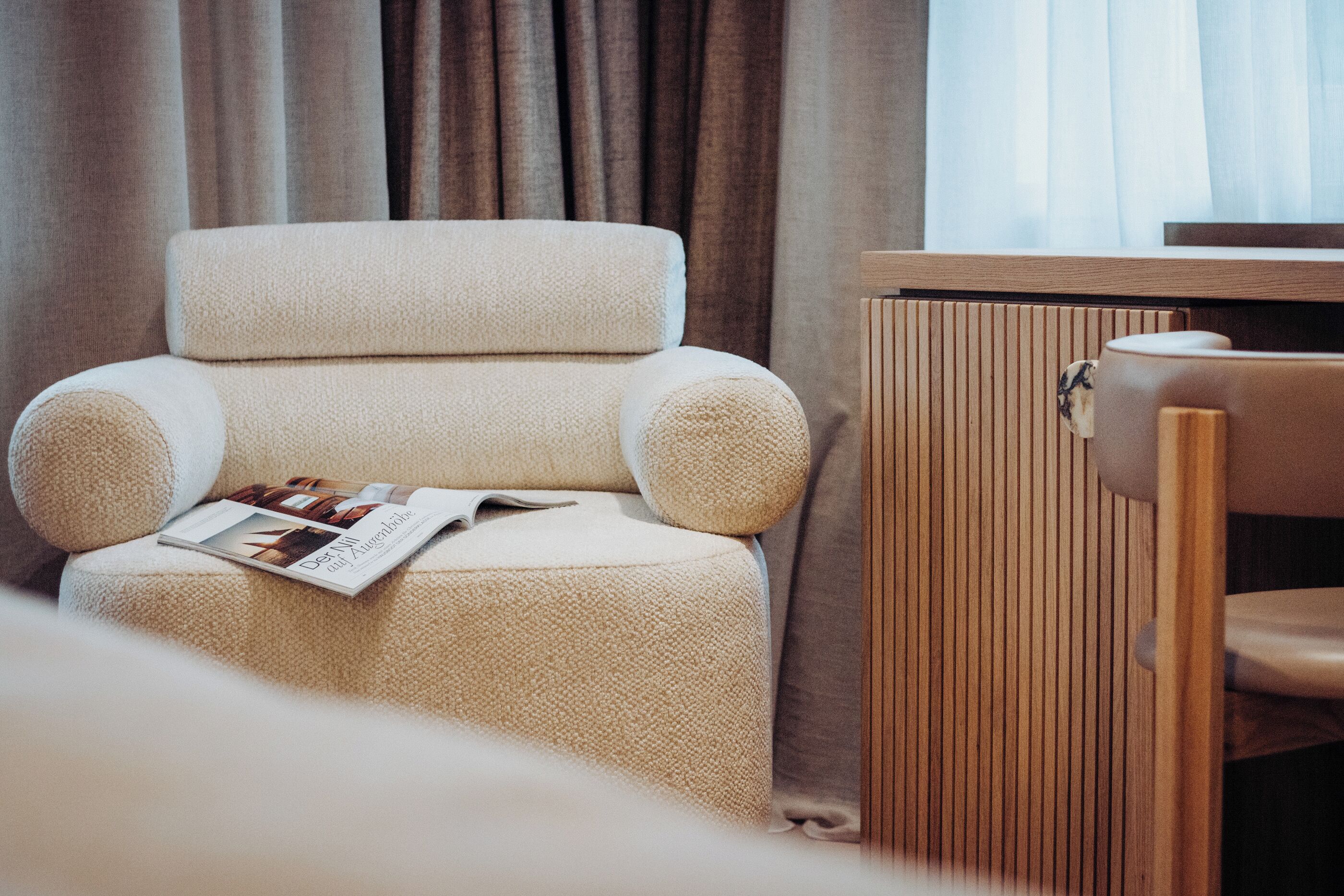 A cozy room with a plush armchair, a wooden side table, and a magazine on the armchair.