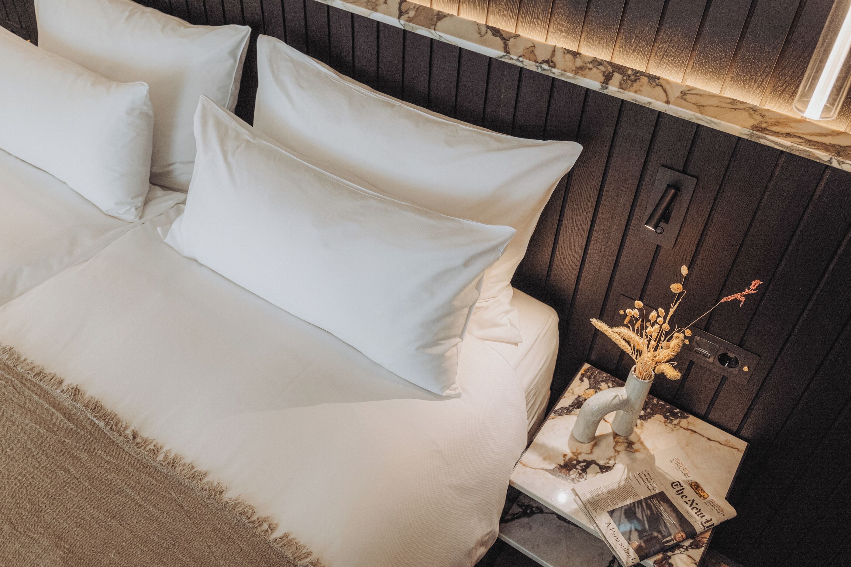 A neatly made bed with white linens and a beige throw blanket. A small bedside table holds a vase with dried flowers and a newspaper.