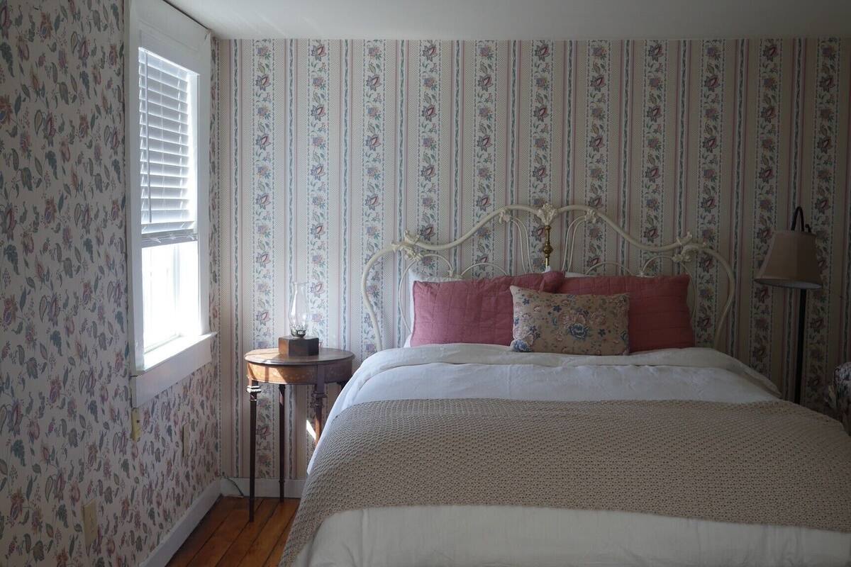 A bedroom with a wooden floor, a bed with a headboard, a bedside table, and a window with blinds.