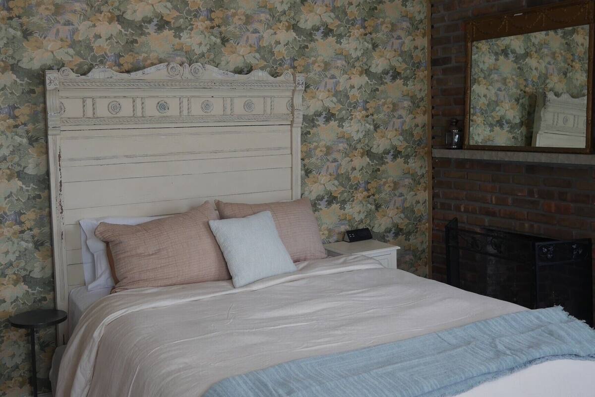 A bedroom with a floral wallpaper, a wooden headboard, a bed with multiple pillows, a nightstand, and a fireplace.