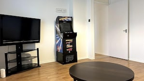 Um quarto com TV de tela plana, máquina de fliperama e mesa redonda.