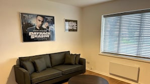 Uma sala de estar com um sofá verde escuro, uma mesa de centro redonda de madeira e dois cartazes de filmes na parede.