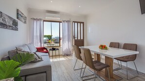 A modern living room with a sofa, a dining table with chairs, a small table with a vase, and a large window with curtains. - Pateo Loft Albufeira (Albufeira)