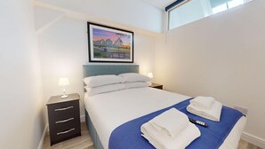 Ein Hotelzimmer mit einem ordentlich bezogenen Bett, einem Rahmenbild mit einer Landschaft darüber, einem Nachttisch mit Lampe und einer blauen Tagesdecke.