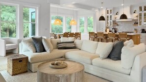 A spacious living room with a large L-shaped white sofa, a round wooden coffee table, and a ceiling fan. - 5 BR Luxury Beach Rental w Pool, Amenities Galore (Isle of Palms)