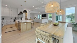A modern kitchen with white cabinetry, a central island, and a dining area with a large wooden table and wicker chairs. - 5 BR Luxury Beach Rental w Pool, Amenities Galore (Isle of Palms)