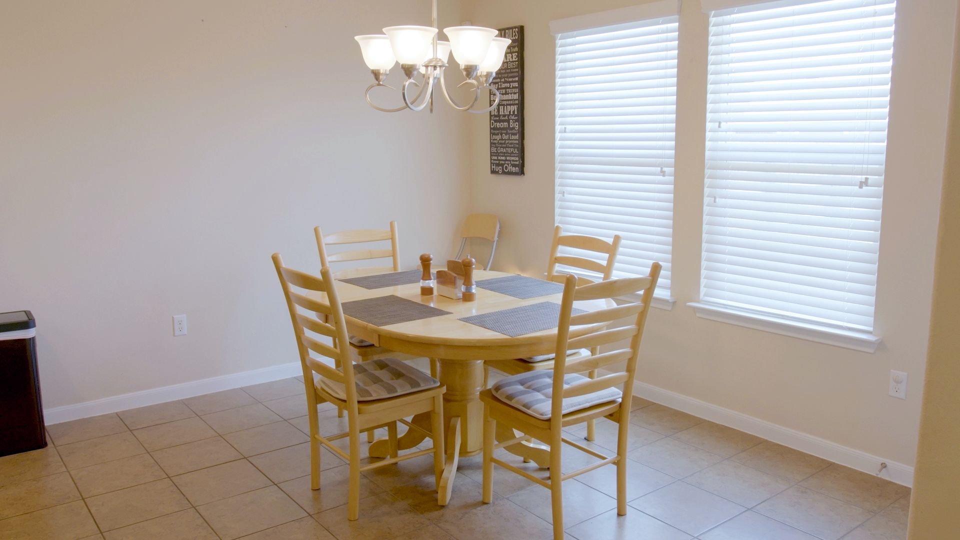 Un coin repas comprenant une table ronde, six chaises, un lustre et une fenêtre avec des stores.