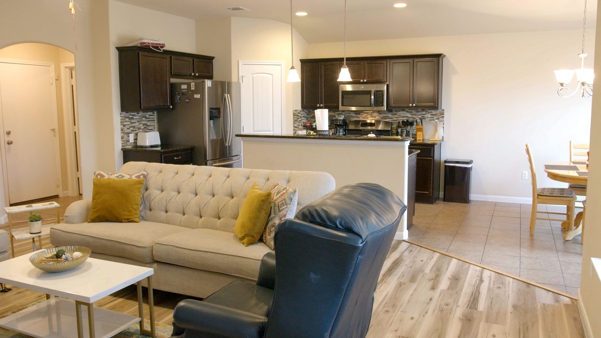 A modern living room with a sofa, a dining table, and a kitchen area with dark cabinets and stainless steel appliances.