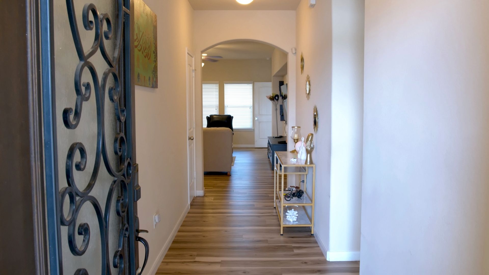 Un couloir doté d’un plancher en bois, d’une grille décorative en métal et d’une porte blanche donnant sur une pièce où se trouvent un canapé blanc et une petite table.