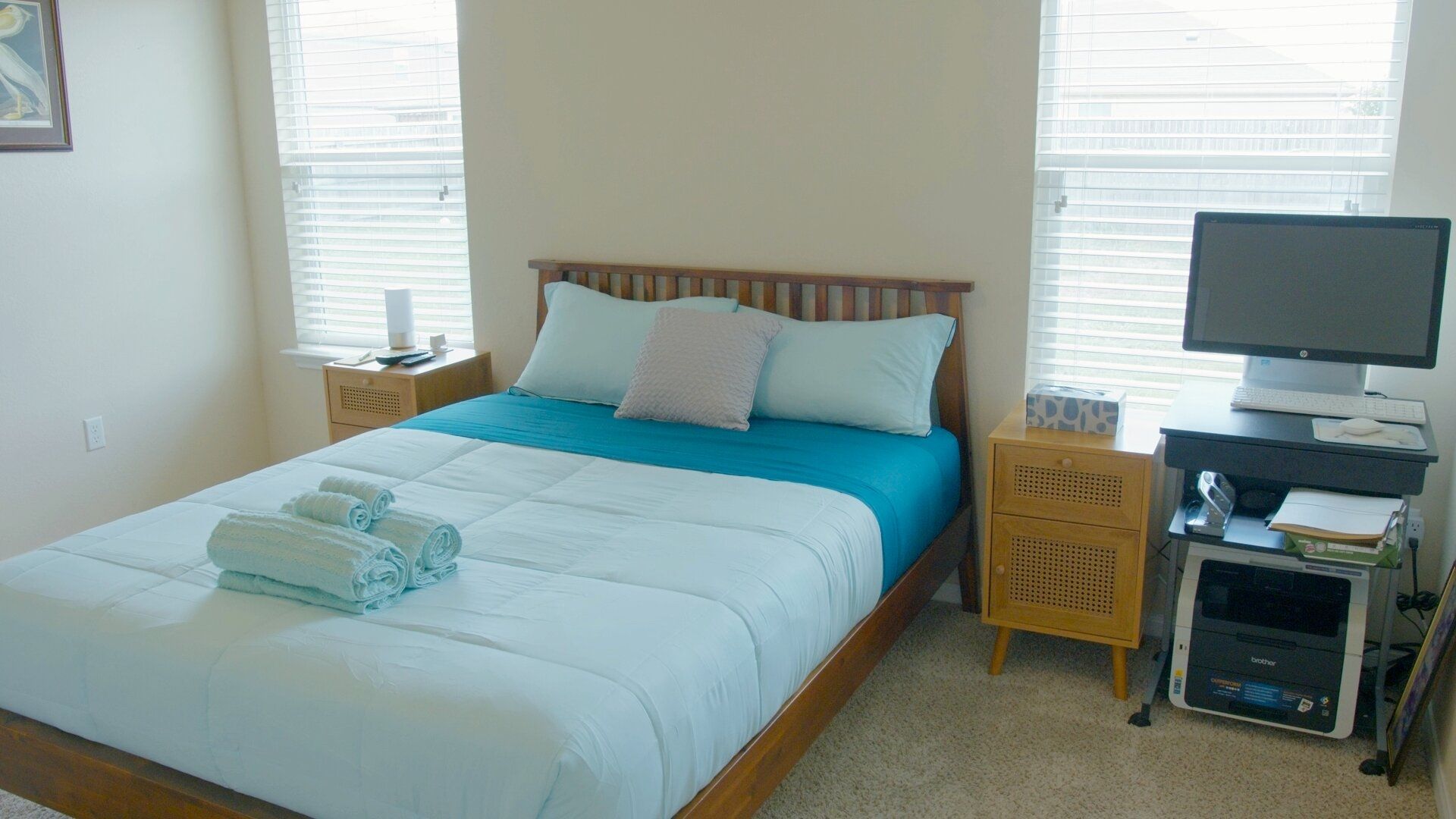 Una cama bien hecha con ropa de cama azul y almohadas blancas, una mesita de noche con una lámpara, y un escritorio con una computadora e impresora.