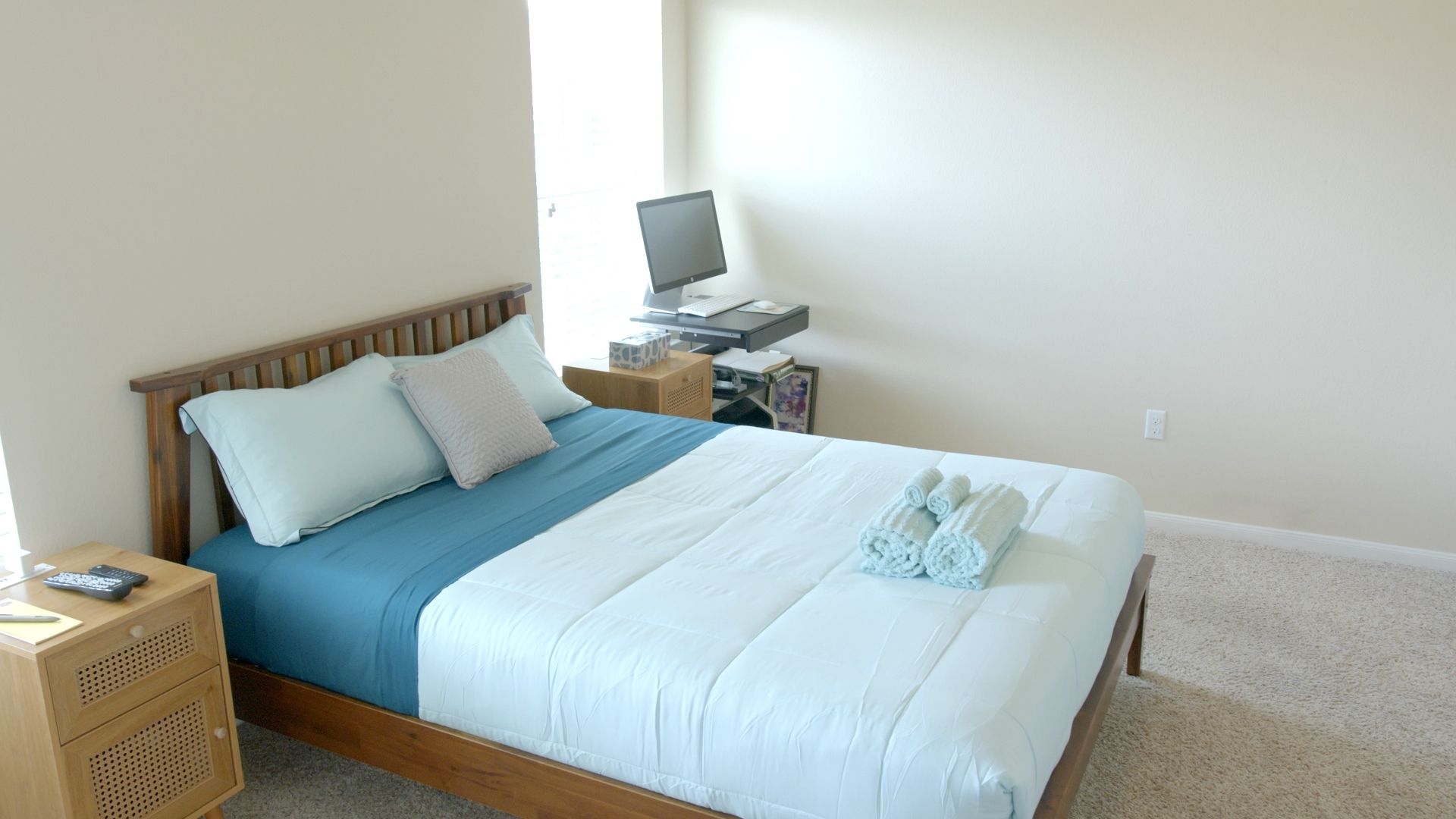 Un lit bien fait, avec une couette bleue et blanche, une tête de lit en bois et deux oreillers. On aperçoit une petite table de chevet avec une télécommande et un livre. Un bureau avec un ordinateur et une lampe est visible à l’arrière-plan.