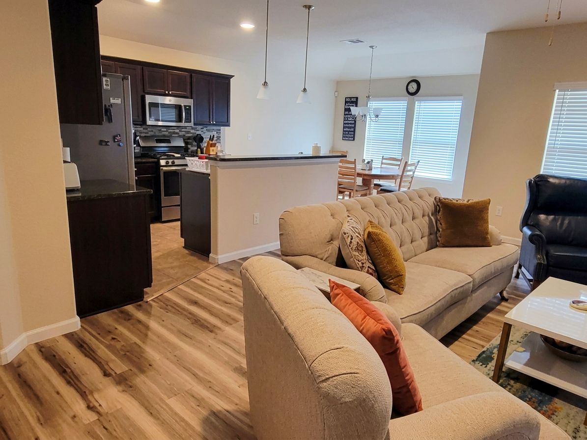 Een moderne keuken met donkere kastjes, een magnetron en een centraal eiland. Er staat een beige bank met een kussen en een zwarte leren fauteuil. In de achtergrond is een eettafel met stoelen te zien.