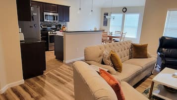 Een moderne keuken met donkere kastjes, een magnetron en een centraal eiland. Er staat een beige bank met een kussen en een zwarte leren fauteuil. In de achtergrond is een eettafel met stoelen te zien.