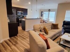A modern kitchen with dark cabinets, a microwave, and a central island. There is a beige sofa with a cushion and a black leather chair. A dining table with chairs is visible in the background.