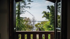 A view through a doorway with a wooden railing, leading to a lush green landscape with trees and a body of water in the distance.