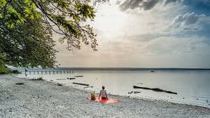 A person sitting on a red mat on a pebbled beach with a calm body of water and a distant shoreline.