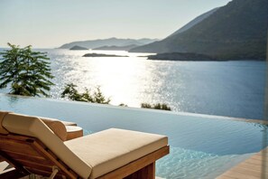 An infinity pool with a wooden deck and lounge chairs, overlooking a calm body of water and distant mountains. - Ficus Seafront Villa, Eco Pool & Close to Beach (CQ6M+WCC Bali, Greece)