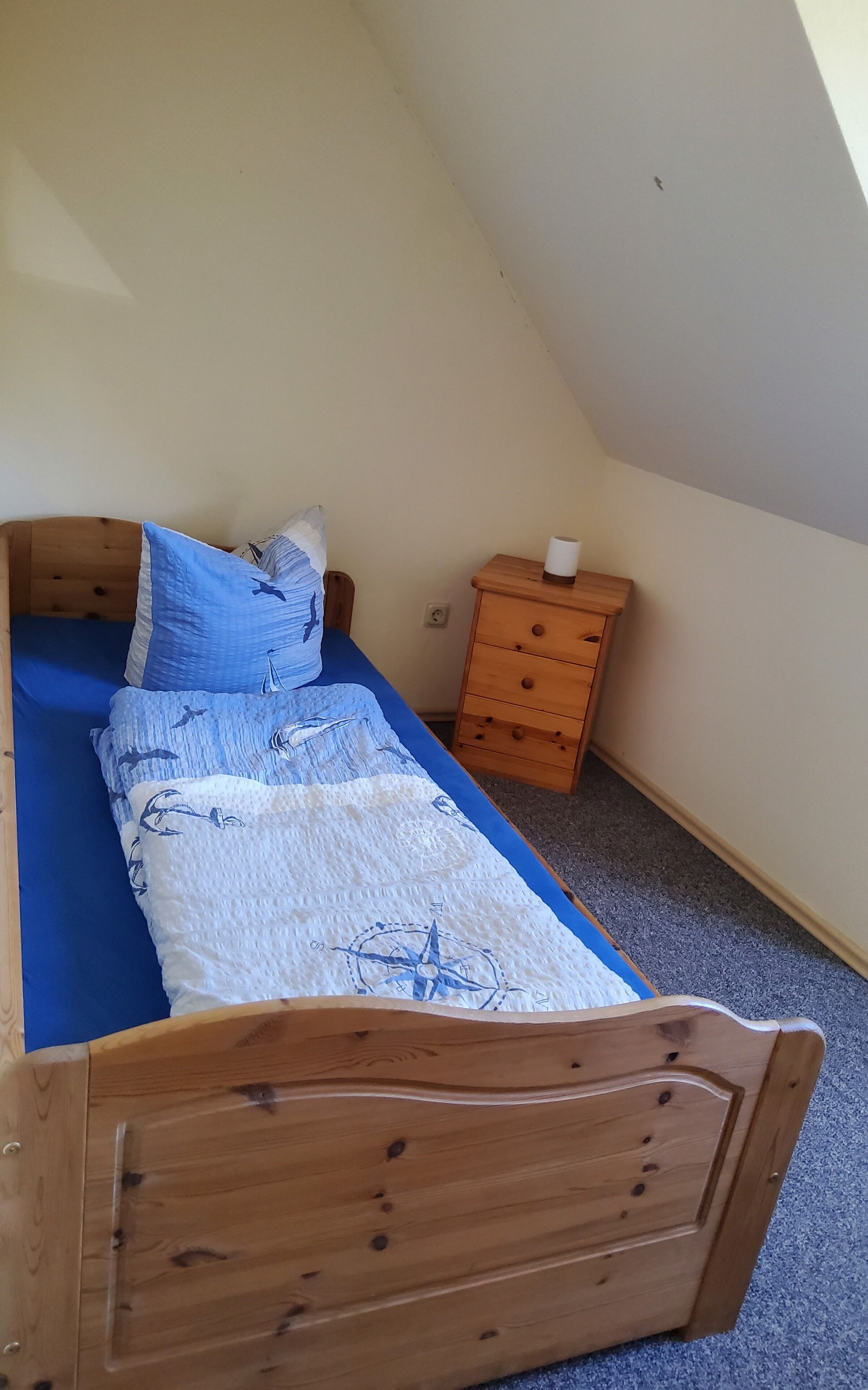 A small, single-bed room with a wooden headboard, a blue bedspread, and a wooden nightstand with a lamp.