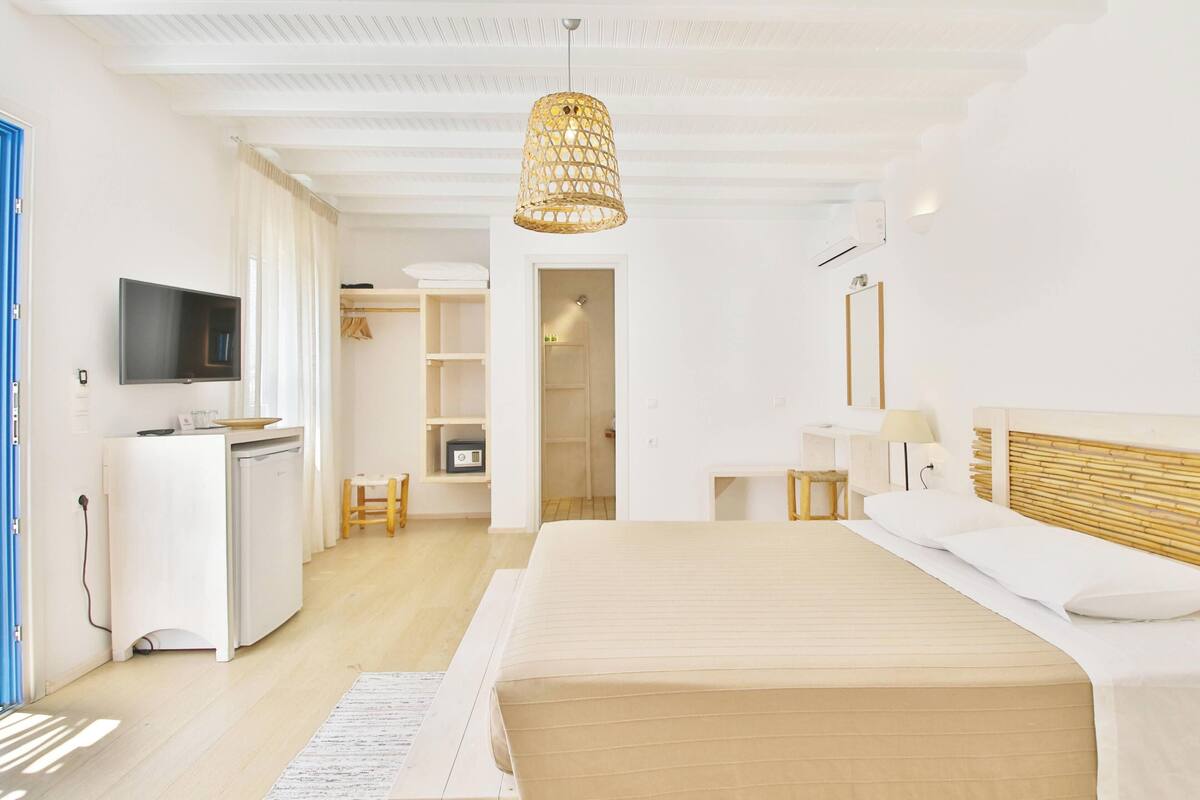 A modern hotel room with a bed, bedside table, lamp, TV, wardrobe, and a kitchenette.