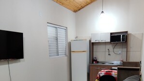 A modern living space with a wooden ceiling, a flat-screen TV, a sofa, a kitchen area with a microwave, and a window with blinds.