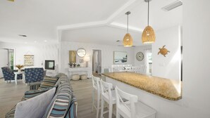 Een moderne woonkamer met een gestreept bankje, witte stoelen, een keukenblok en hanglampen.