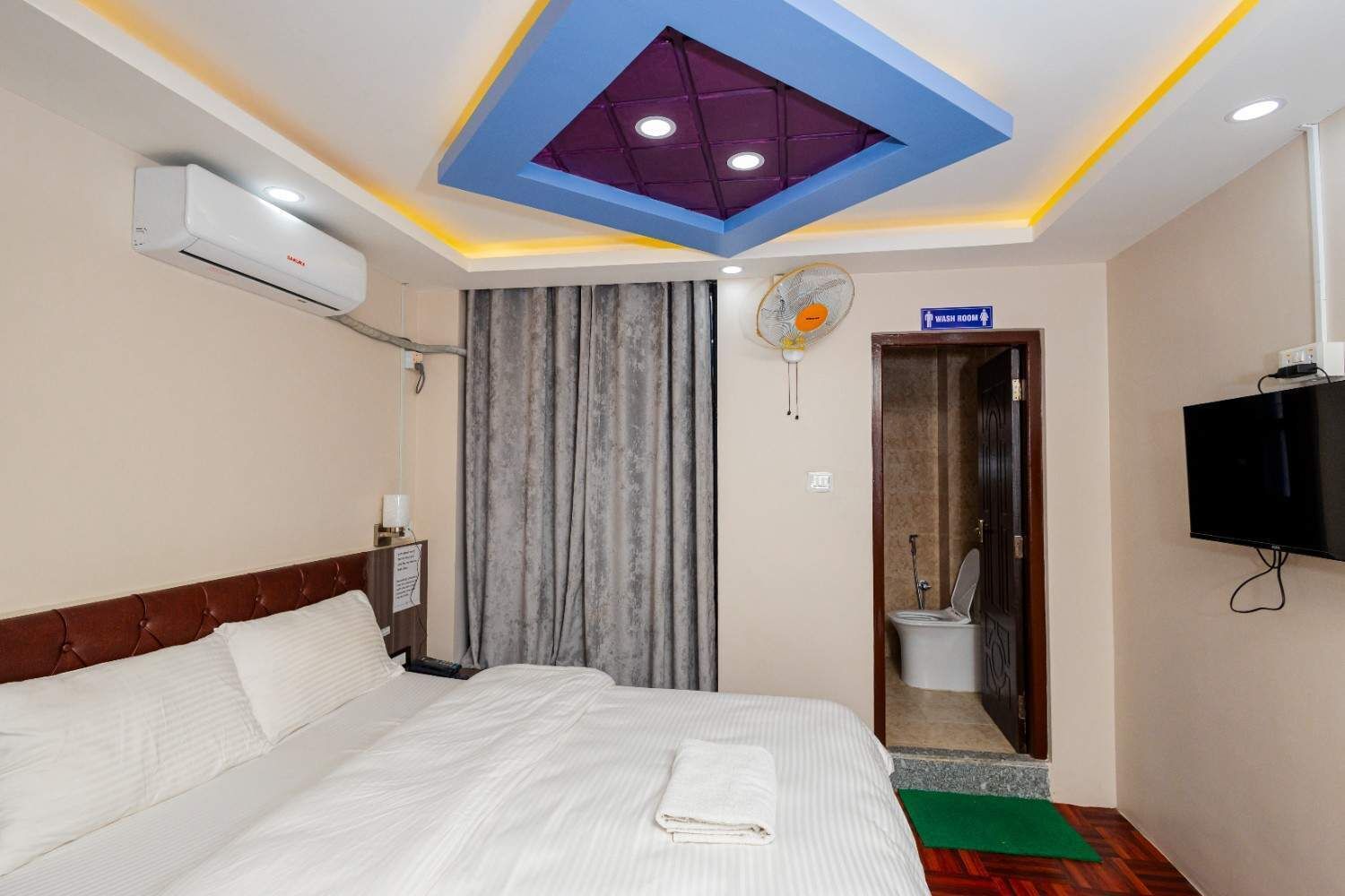 Habitación de hotel con cama, aire acondicionado, ventilador de techo, televisor y un letrero que indica un baño.