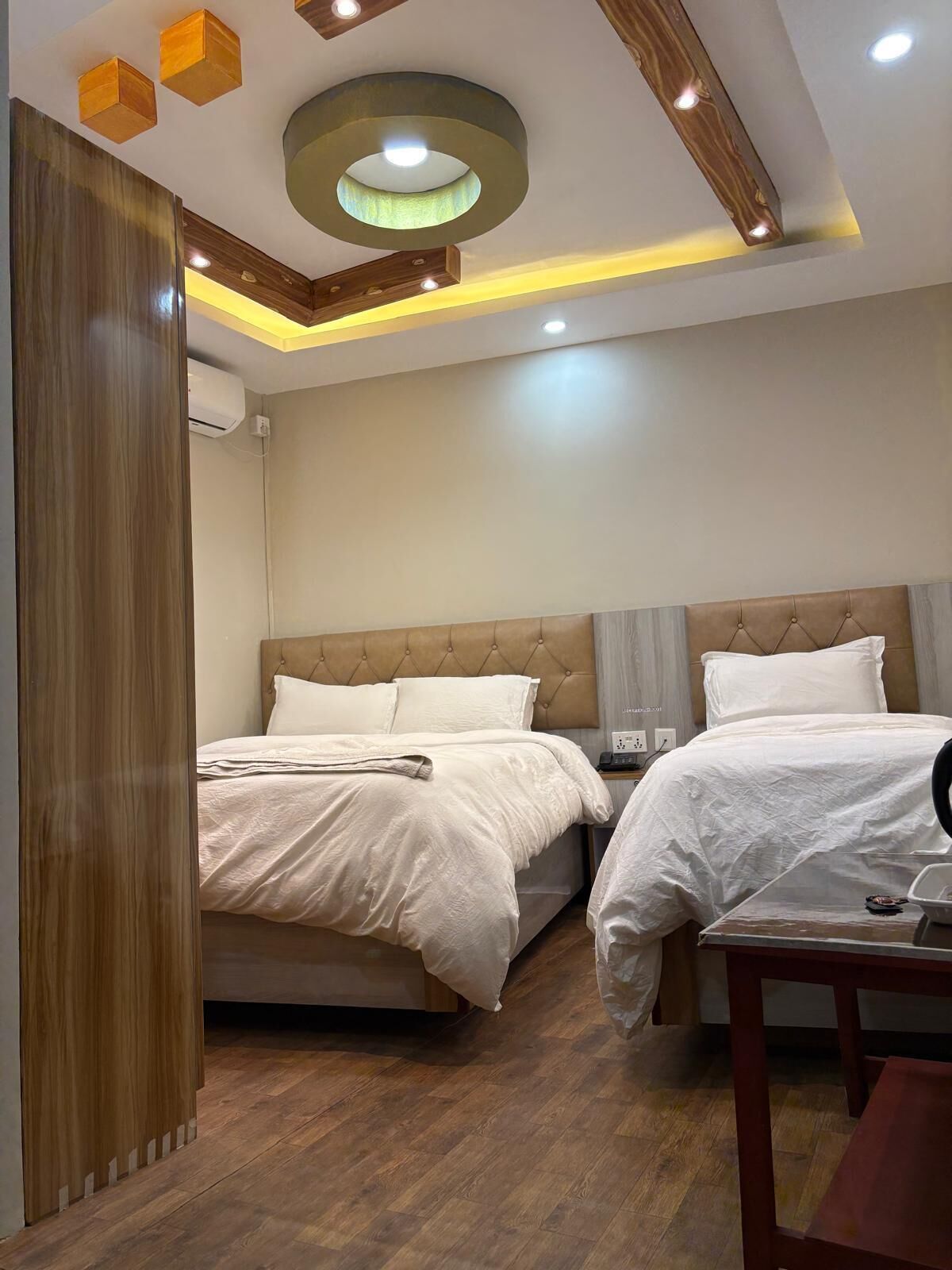 Habitación de hotel con dos camas, suelo de madera, diseño moderno en el techo y una mesita de noche con lámpara.