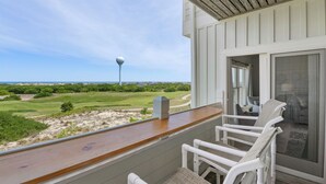 A balcony with a view of a golf course and a lighthouse, featuring white wicker chairs and a wooden deck. - Serenitee: Ocean & Golf Course Views, Shared Pool (Corolla)