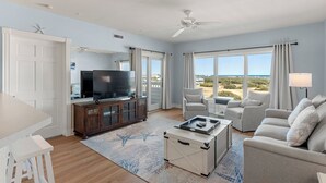 A spacious living room with a television, a sofa, a coffee table, and a view of the beach. - Serenitee: Ocean & Golf Course Views, Shared Pool (Corolla)