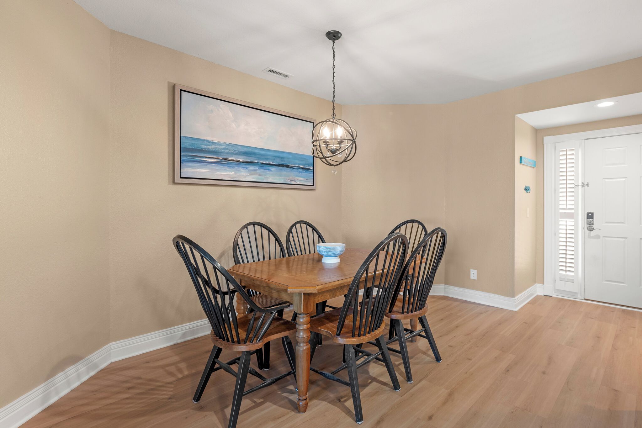 A dining area with a wooden table and chairs, a hanging light fixture, a framed picture on the wall, and a white door in the background.