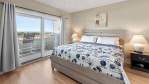 A bedroom with a large bed, a ceiling fan, a view through a sliding glass door, and a nightstand with a lamp. - Serenitee: Ocean & Golf Course Views, Shared Pool (Corolla)