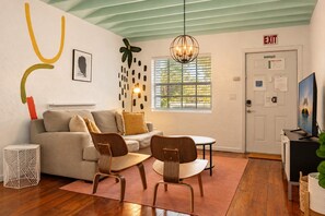Un salon moderne comprenant un canapé, deux fauteuils en bois, une petite table, un lustre, une porte avec un panneau indiquant la sortie, et une fenêtre avec des stores.