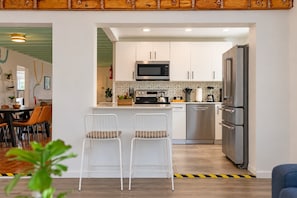 A modern kitchen with white cabinets, a stainless steel refrigerator, and a microwave. There are two white bar stools and a dining table with chairs.
