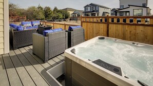 Una terraza de madera con un jacuzzi, mobiliario de mimbre y vistas a casas al fondo.