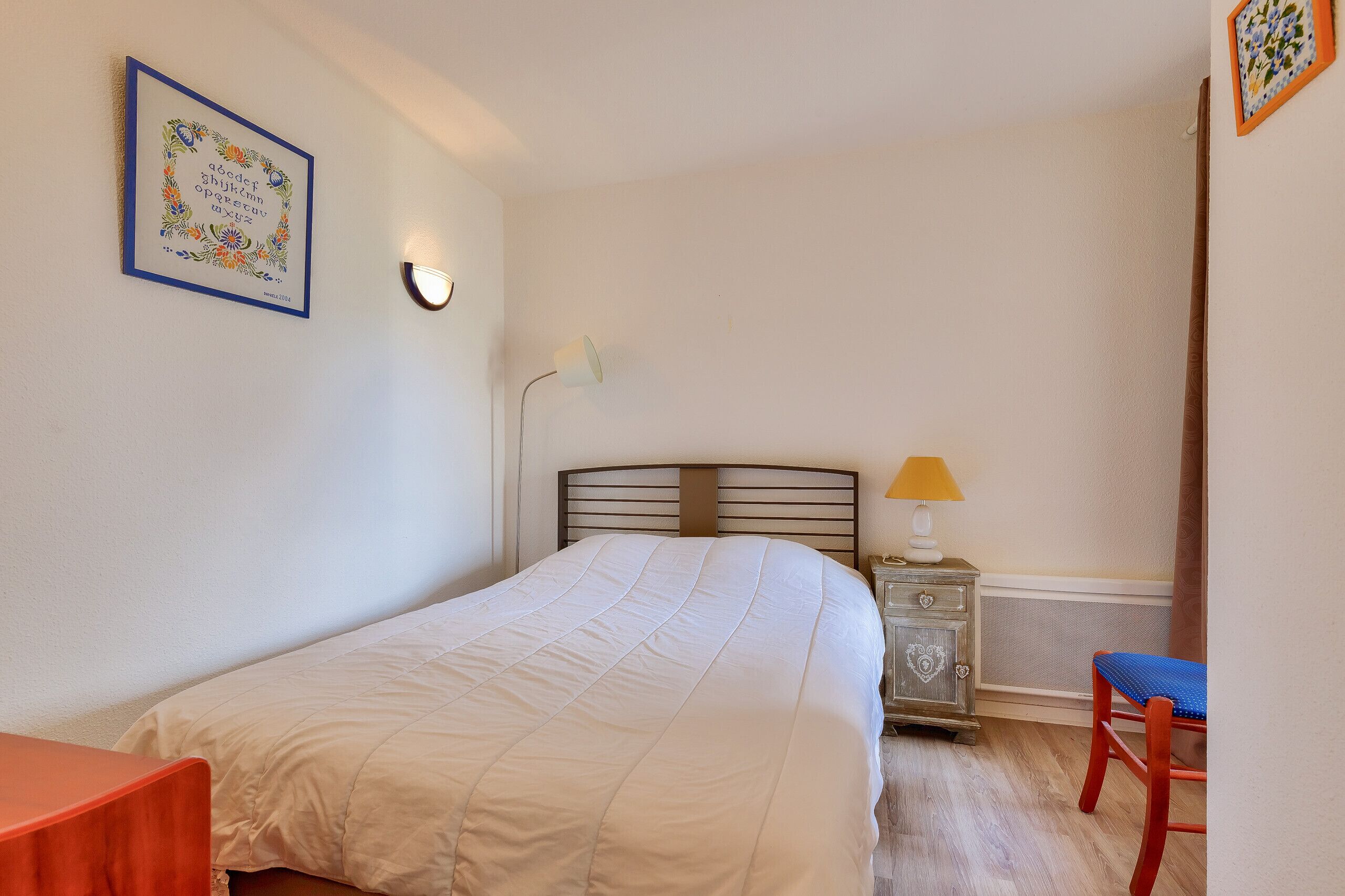 Un dormitorio con cama, mesita de noche, silla, lámpara y un cuadro en la pared.