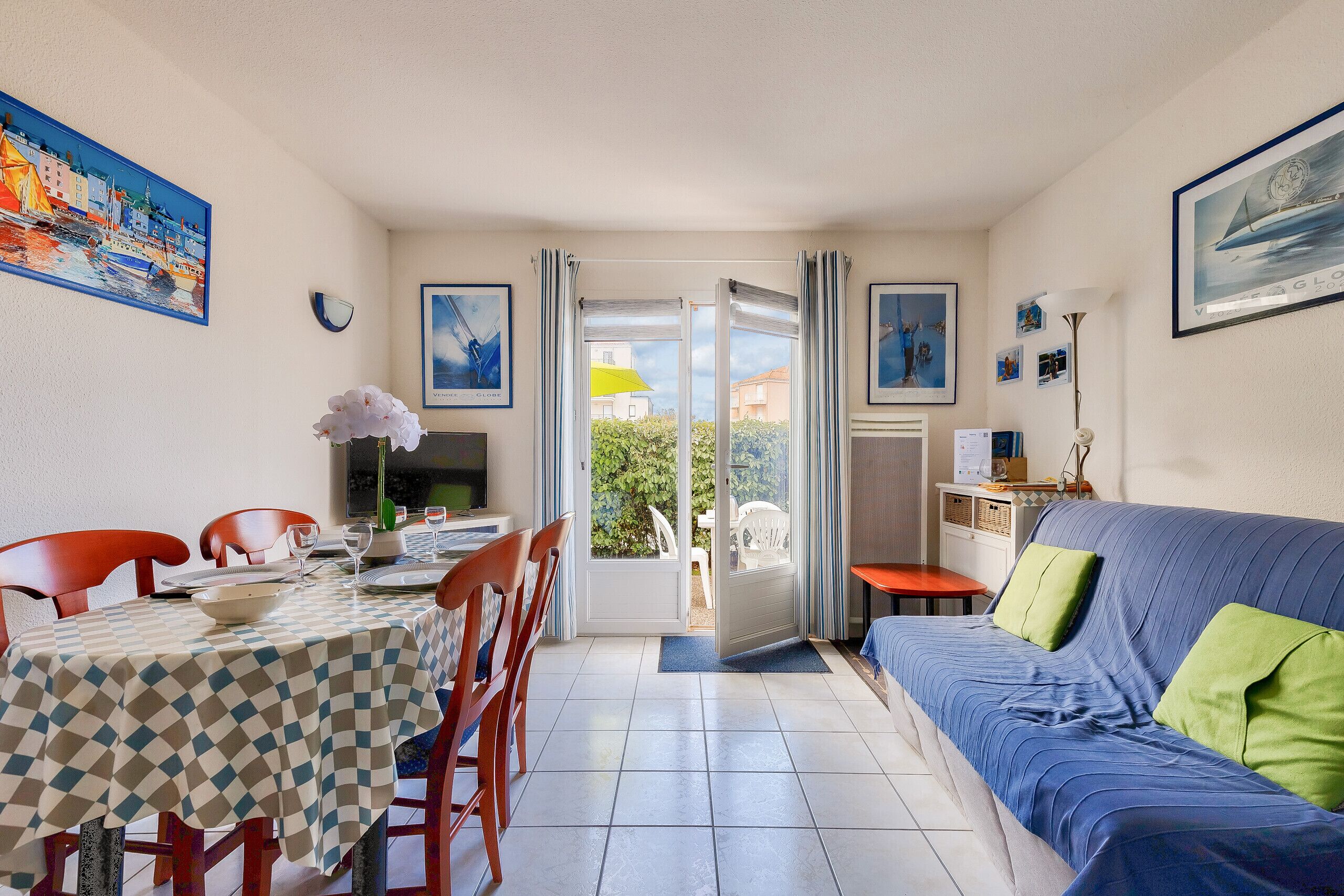 Un comedor con mantel a cuadros, un sofá con cojines verdes y vistas a una zona de estar exterior a través de una puerta de cristal.