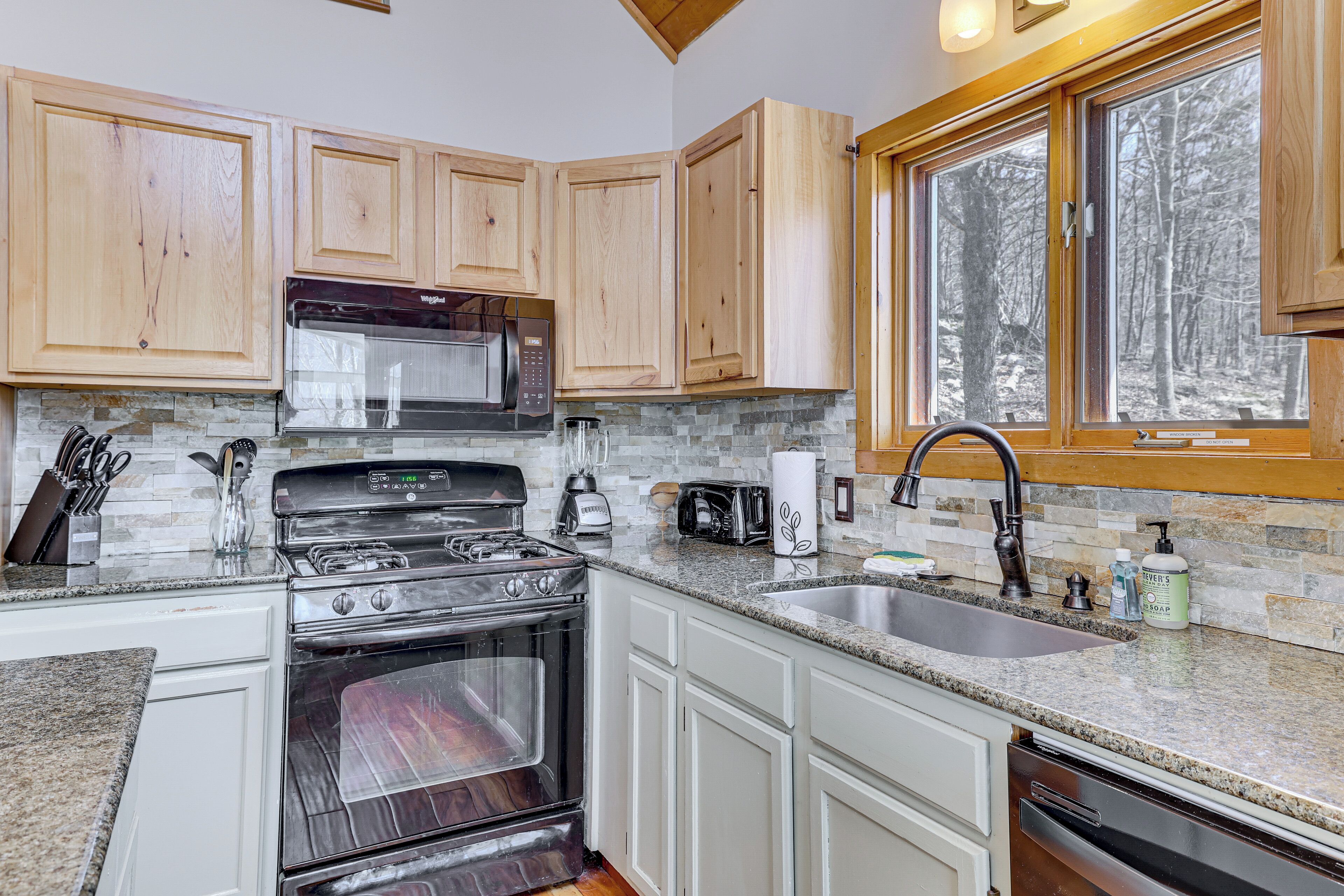 A kitchen with wooden cabinets, a granite countertop, a built-in oven, a microwave, and a window with a view of trees.