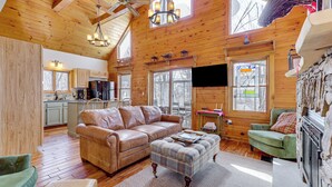 A spacious living area with a wooden ceiling, a large sofa, a plaid ottoman, and a stone fireplace. - Jeffersonville Home w/ Deck Near Hiking & Climbing (Jeffersonville)