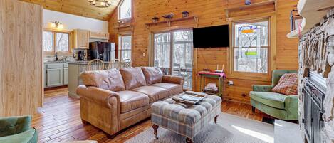 A spacious living area with a wooden ceiling, a large sofa, a plaid ottoman, and a stone fireplace.