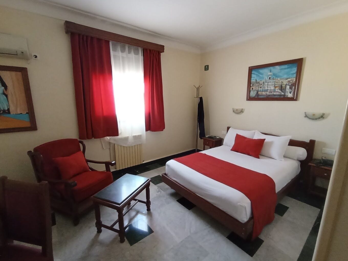 Habitació d'hotel amb un llit, una cadira, una taula petita, una finestra amb cortines vermelles i un quadre a la paret.