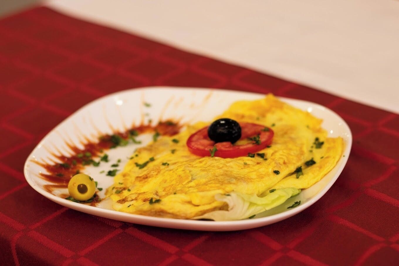 A plate with an omelette, a slice of tomato, a black olive, and a garnish of herbs.