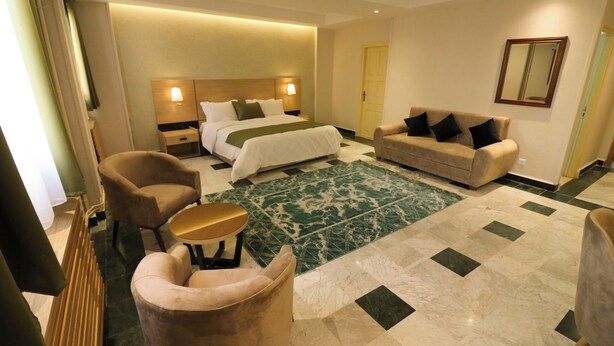 Una habitación de hotel con una cama, dos sillones, un sofá, una mesita redonda y un tapete verde estampado.