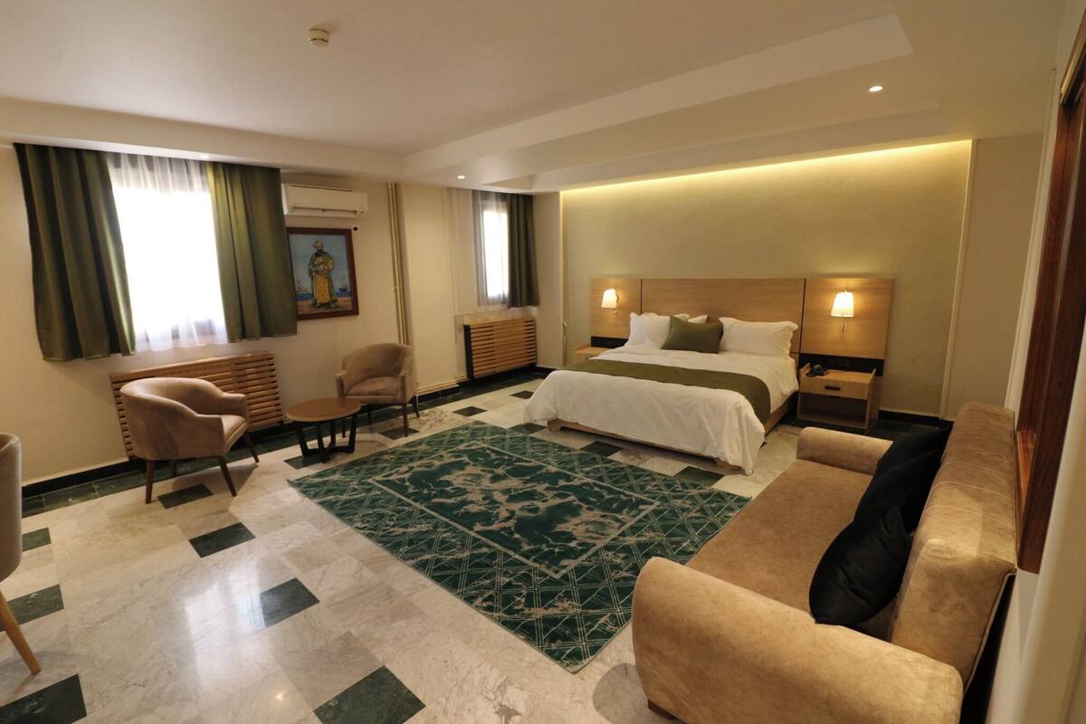 Una habitación de hotel con cama, dos sillones, un sofá, alfombra verde estampada, cabecera de madera, lámparas de noche y un cuadro en la pared.
