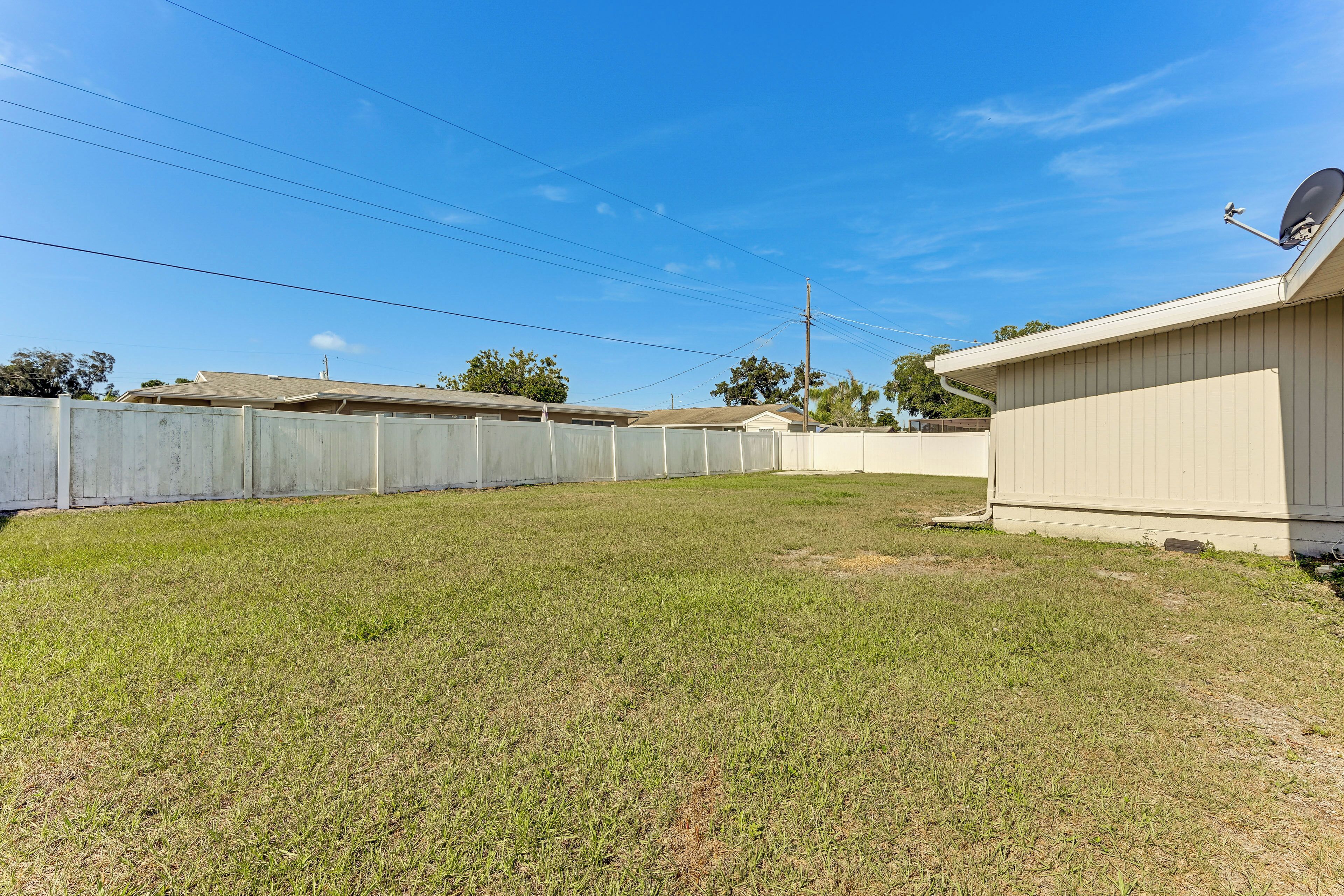 A fenced backyard with a grassy lawn, a building with a satellite dish, and a clear blue sky.
