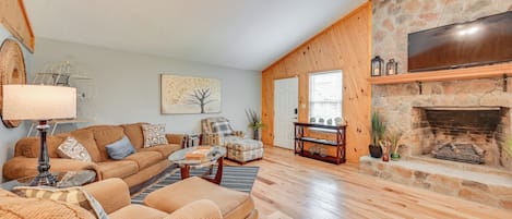 A cozy living room with a stone fireplace, a large TV mounted above it, and a comfortable seating area with a sofa and armchairs.